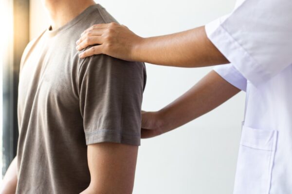 Fysioterapeut undersöker manlig patient i grå t-shirt med rygg och axel-problem.