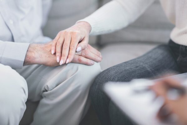 Äldre man hos samtalsterapeut. En kvinna i jeans och med rosa gelenaglar lägger sin hand på mannens hand. I förgrunden för samtalsterapeuten anteckningar.