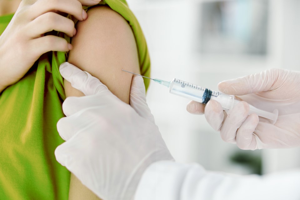 Patient med grön tröja får vaccin injicerat i överarmen av vårdpersonal med vita plasthandskar.